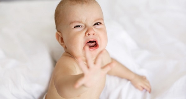 Qu'est-ce qu'une quantité normale de pleurs: photo d'un bébé qui pleure, assis sur un drap blanc et tend la main vers l'avant.