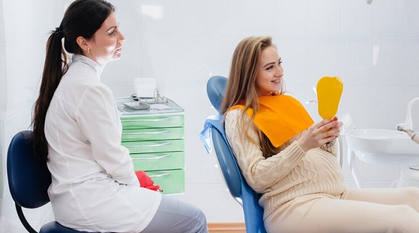 Pourquoi une visite chez le dentiste pendant la grossesse peut contribuer à la santé de la mère et du bébé: photo d'une femme enceinte tenant un miroir jaune assise devant un medecin.